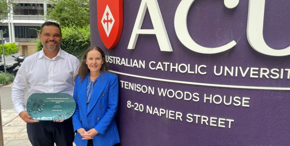 An Aboriginal man in a white shirt and a white woman in a blue shirt stand next to a sign for Australian Catholic University. The man is holding an award from Indigenous Allied Health Australia.