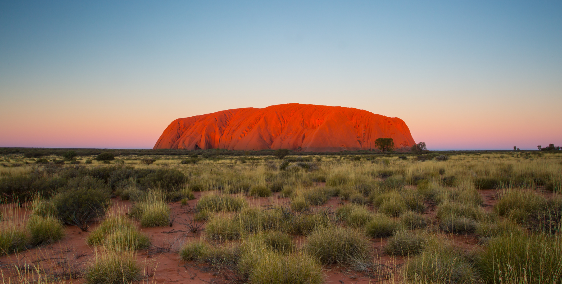 Photo of Uluru with colourful sky behind it, indicating the sun is rising